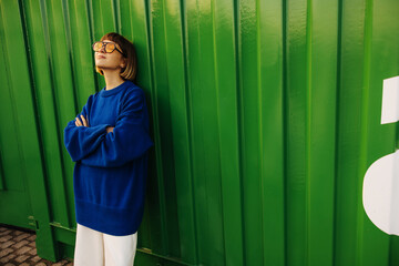 Nice young caucasian girl eyes closed stands leaning against green wall with space for text. Brown-haired woman wears sunglasses, blue sweatshirt and white pants. Concept of enjoying moment