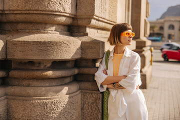 Wall Mural - Stylish young caucasian woman looking away, posing with her arms crossed outdoors. Short, brown-haired wears casual clothes and sunglasses in summer. City life concept