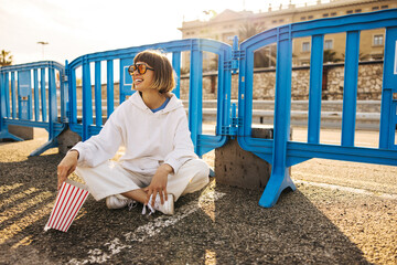 Wall Mural - Cheerful young caucasian lady enjoys spending time outdoors sitting on pavement. Brown-hair with bob haircut wears white hoodie and pants in sunny weather spring. Lifestyle concept