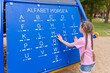 A little girl 7-8 years old with long hair dressed in a pink T-shirt and shorts is studying Morse code on a blackboard in the playground.