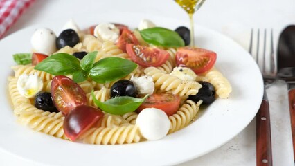 Wall Mural - Pouring olive oil on cold summer pasta salad with black olives, cherry tomatoes and mozzarella cheese. Closeup view