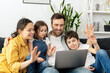 © Vadim Pastuh - Virtual meeting. Family of four waving to screen while using laptop for video connection with grandparents or family. Multiracial mom, dad and two kids waving in webcam of the laptop, video call