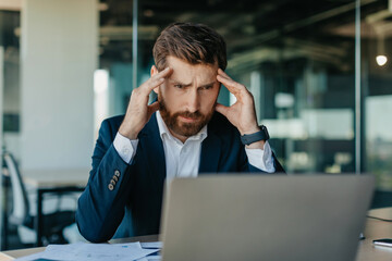 Depressed middle aged businessman looking at laptop and touching head, suffering from headache or having problems