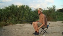 A Man Fisherman At Sunset In A Hat On The Lake Catches Fish With A Fishing Rod Against The Background Of Reeds. Sits On A Chair Kills Mosquitoes. Fishing Holiday Concept. Harmful Mosquitoes. C