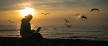 Caucasian Woman Typing On A Laptop On The Seashore At Sunset. Freelance Work. Wide Screen. 