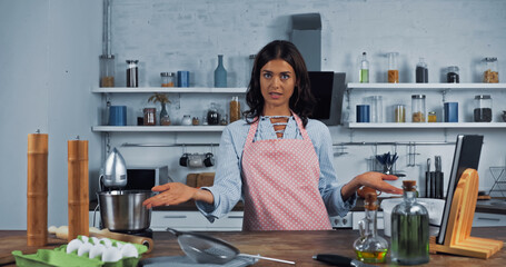 Wall Mural - culinary vlogger pointing at ingredients and cooking utensils while looking at camera.