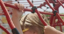 Blond Boy In Sunglasses Plays On Children Playground. Preschooler Climbs On Red Climbing Rope Net. Child Has Fun Outdoors On Sunny Day Closeup