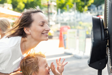 Profile side close-up view young adult woman with son enjoy fresh moisturized air blowing from big cooling fan machine hot sunny summer weather outdoors. Mist spraying ventilator system city street