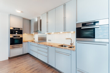 Large luxury kitchen unit made of light blue laminate, glass and marble. It is equipped with premium built-in appliances. The kitchen is lit by warm spot LED lights. The floor is wooden.