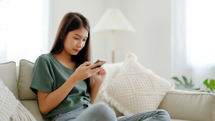 Happy young asian woman relax on comfortable couch at home texting messaging on smartphone, smiling girl use cellphone chatting, browse wireless internet on gadget, shopping online from home