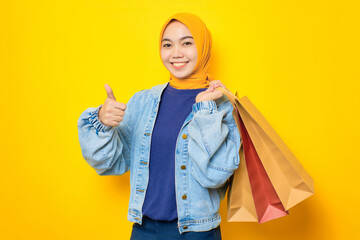 Happy young Asian woman in jeans jacket holding shopping bags and gesturing thumbs up, recommending great sale isolated over yellow background
