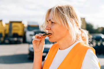 Wall Mural - Middle aged portrait of worker engineer woman with orange vest eating cookies. Female truck driver 