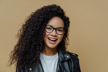 Headshot of cheerful African American woman with dark curly hair, has pleased face expression, joyful conversation, wears transparent glasses and black leather jacket, isolated on beige background