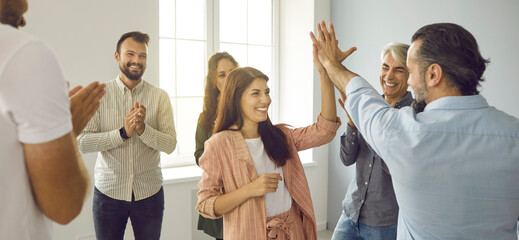 Wall Mural - Teamwork concept. Header background with team of cheerful people having fun workshop with business coach. Happy positive man and woman giving each other high five standing in office among coworkers