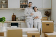 © VK Studio - Smiling relaxed husband and wife drink coffee at kitchen, hold paper cups with hot beverage, dressed in casual outfit, relocate in new apartment, pile of cardboard boxes with property in foreground.