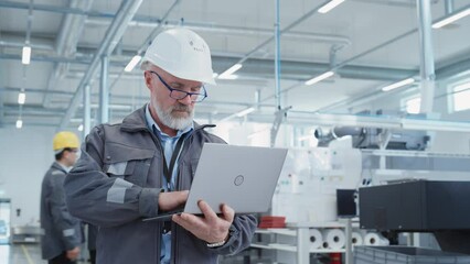 Fototapete - Portrait of a Bearded Middle Aged General Manager Standing in a Factory Facility, Wearing Work Jacket and a White Hard Hat. Heavy Industry Specialist Working on Laptop Computer.
