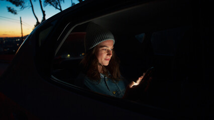 Side night cinematic view of young beautiful woman sit in taxi car back seat near window on road trip, hold, check on mobile phone, use smartphone, scroll social media news, read messages and smile