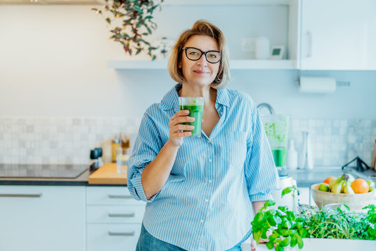 Smiling middle aged woman with just made glass of detox shake, green smoothie in the kitchen. Healthy dieting, eating, cooking. Natural anti aging methods, weight loss program. Vegan, vegetarian diet.
