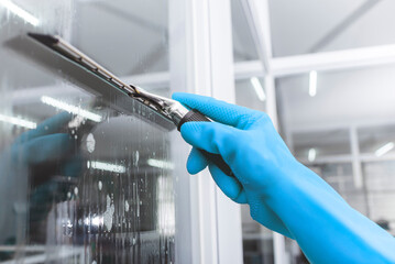 A man wearing light blue rubber gloves cleans the surface of an interior office window with a glass wiper or squeegee. The surface is sprayed with soap.