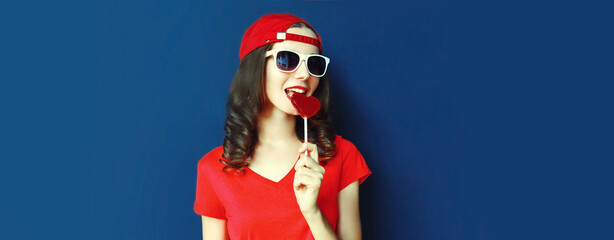 Portrait of young woman with heart shaped red lollipop wearing baseball cap, sunglasses on blue background