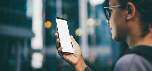 young woman looks at phone and walking down street against skyscrapers in evening twilight backside 