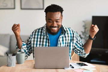 Wall Mural - Joyful African American Guy Using Laptop Shaking Fists Sitting Indoors