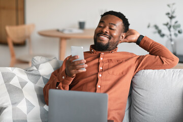 Wall Mural - Cheerful African American Guy Using Smartphone And Laptop At Home