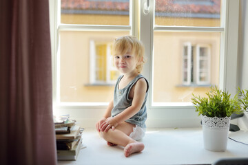 Cute toddler boy standing on a windowsill. Adorable child looking out the window.