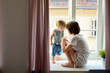 Teenage sister and her toddler brother looking out the window. Siblings sitting on a windowsill.