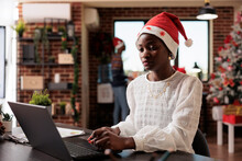 Company Employee Using Laptop At Office Job, Celebrating Winter Season With Christmas Decorations And Tree. Woman With Santa Hat Working In Space With Festive Seasonal Ornaments.