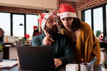 Festive People Doing Teamwork In Company Office With Christmas Tree And Lights, Using Laptop To Work On Business. Man And Woman With Santa Hat At Job With Holiday Decorations And Ornaments.