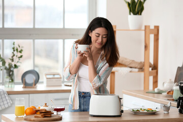 Poster - Beautiful young Asian woman drinking coffee and making tasty toasts in kitchen