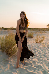 Wall Mural - Cheerful brunette woman in stylish summer  boho outfit posing on the beach.  Sunset colors.