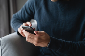 Happy Asian man using smartphone on sofa in living room at home, searching data and social media on internet.