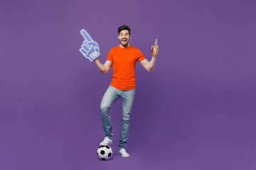 Wall Mural - Full body young fan man he wear orange t-shirt cheer up support football sport team hold soccer ball fan foam glove finger up beer bottle watch tv live stream isolated on plain dark purple background.