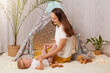 © sementsova321 - Indoor shot of young mother playing with with baby daughter on floor near kids wigwam, expressing positive emotions, spending time with cute kid in peetee tent at home.