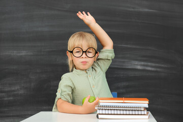 Back to school. Happy smiling pupil in round glasses sitting at the desk. Child raises his hand up in the classroom with blackboard on background. Kid boy from primary school. First day of fall