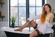 © Denis Kadatsky - a woman with long red hair in a white shirt and stockings sits on a bathtub in the bathroom
