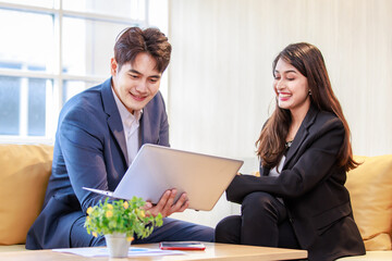 Millennial Asian professional successful male businessman and female businesswoman in formal suit sitting on sofa smiling discussing together while working with laptop notebook computer in office