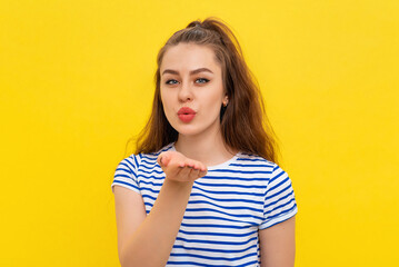 Wall Mural - Pretty young woman sends air kiss tempting perfect lips, flirting boyfriend, stands in white-blue striped t shirt over yellow background