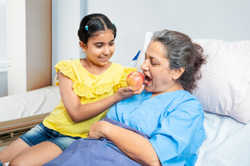 Wall Mural - Little granddaughter help her grandmother patient eating apple in hospital bed. 