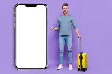 Poster - Photo of positive excited guy wear grey shirt holding suitcase pointing modern device empty space isolated violet color background