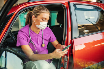 A photo of a young female doctor sitting in her car wearing a protective mask and looking at her phone. A nurse sits in a car and texting on her mobile phone.