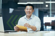 © Liubomir - Portrait of successful Asian investor, man puts money money cash in envelope, smiling and looking at camera, working in modern office
