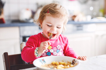 Adorable baby girl eating from fork vegetables and pasta. food, child, feeding and development concept. Cute toddler, daughter with spoon sitting in highchair and learning to eat by itself.