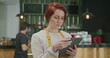 Young employee woman wearing apron standing inside coffee shop holding tablet device checking online orders. Red hair girl working at cafe