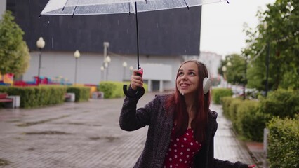 Wall Mural - Young beautiful woman with transparent umbrella dancing in rain on city street. Pretty female in wireless headphones listening to music, sheltering with umbrella from rain on walk in city park.