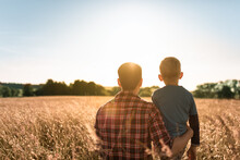 Father And Son Spending Time Together In Nature Relaxing In A Field Looking At The Sunset, Fatherhood, And Family Parenting Concept. 