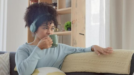 Poster - A woman dancing, listening to music on headphones and drinking a cup of coffee while relaxing on a home living room sofa. Cool afro woman enjoying an off day and sipping a mug of tea during lockdown