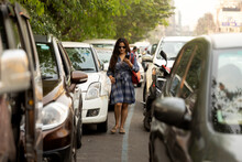 Fashionable Woman Speaking In Smartphone Walking Through Cars In City